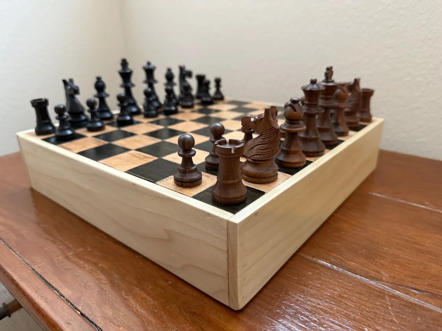 A custom wooden chessboard box with a built-in drawer, shown with hand-carved chess pieces set up on the board.