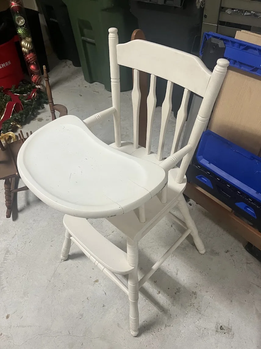 An unrestored wooden baby high chair with worn, peeling white paint and loose joints in a workshop.