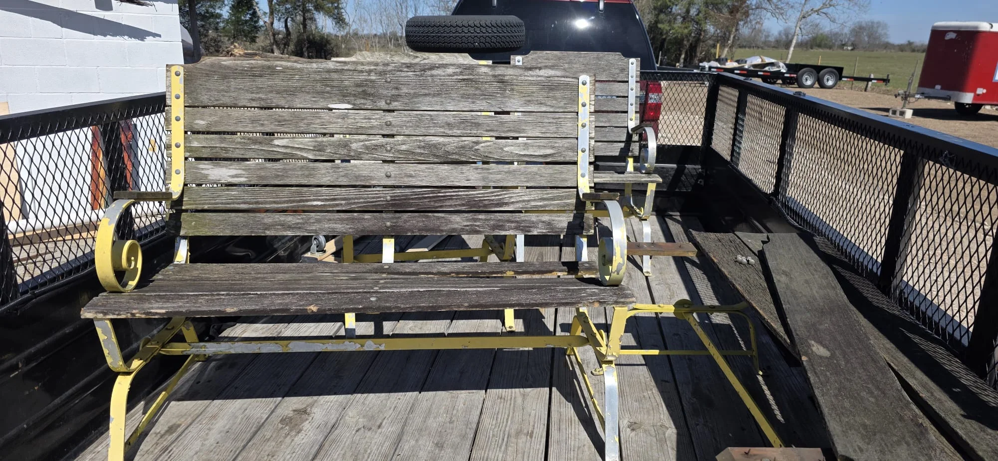 A weathered wooden and metal patio glider bench with peeling yellow paint loaded onto a trailer bed before restoration.