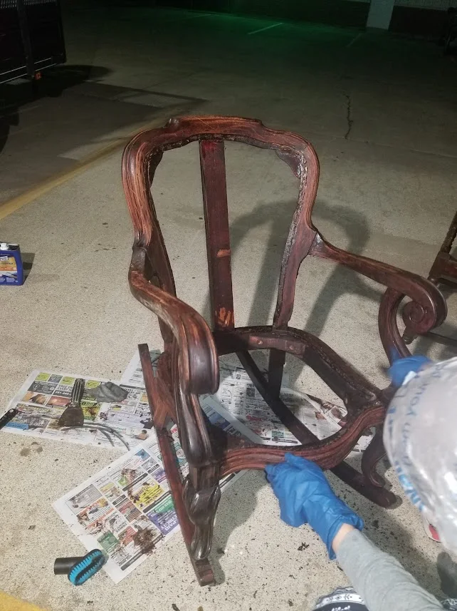 An artisan in blue gloves prepping the stripped wooden rocking chair frame over protective newspaper