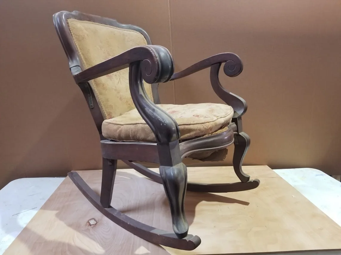 An old wooden rocking chair with faded wood finish and sagging seat cushion on a plywood board